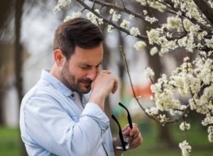 Man standing in front of a flowering tree holding his glasses and sneezing